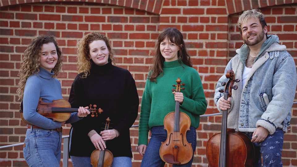 A group of students, wearing long sleeved jumpers, holding their string instruments, smiling at the camera.